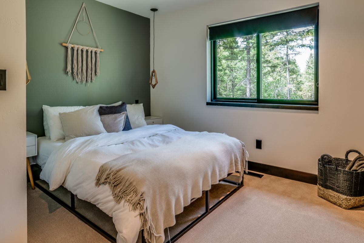 A modern bedroom with a green accent wall featuring a macramé hanging, a bed with white bedding, and a pendant light. A large window provides natural light and a view of trees outside.