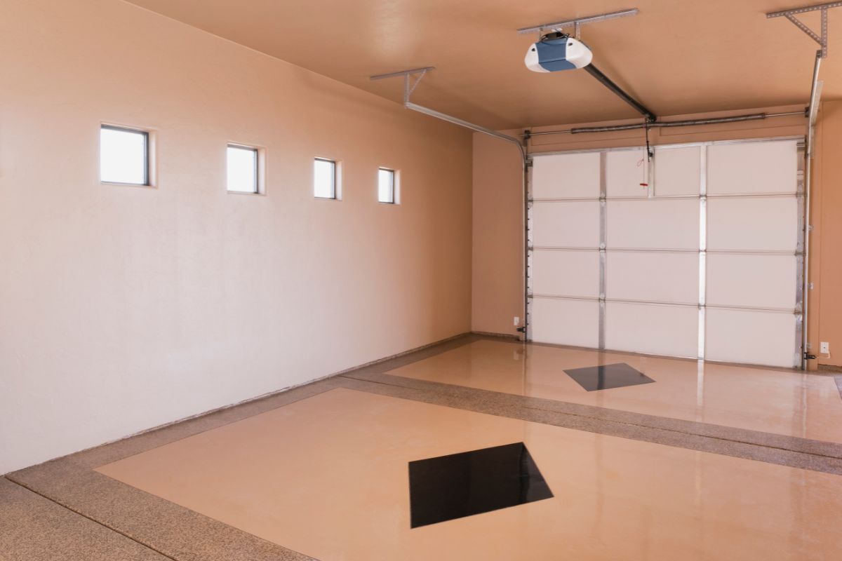 A clean and well-lit garage with smooth, painted walls in a warm peach tone and a glossy epoxy floor. The space features a white roll-up garage door, small windows for natural light, and overhead storage racks, showcasing the results of professional garage and basement painting.