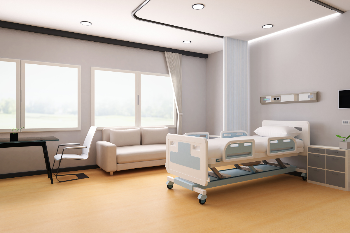 A modern hospital room with neutral paint colors, featuring a patient bed, sofa, and natural light from large windows. The calming color palette enhances the healing environment.