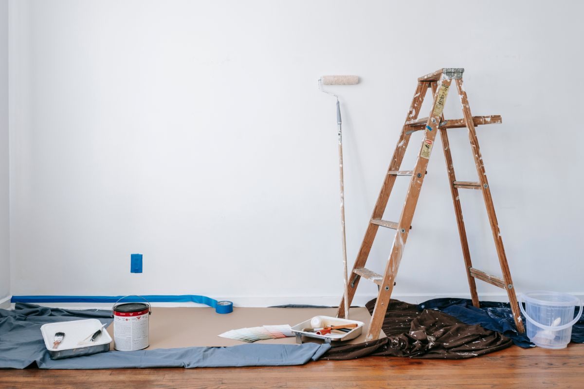 A room prepared for interior painting, featuring a wooden ladder leaning against the wall, a paint roller resting on it, and various painting supplies including a paint can, tray, brushes, tape, and protective drop cloths spread across the floor.