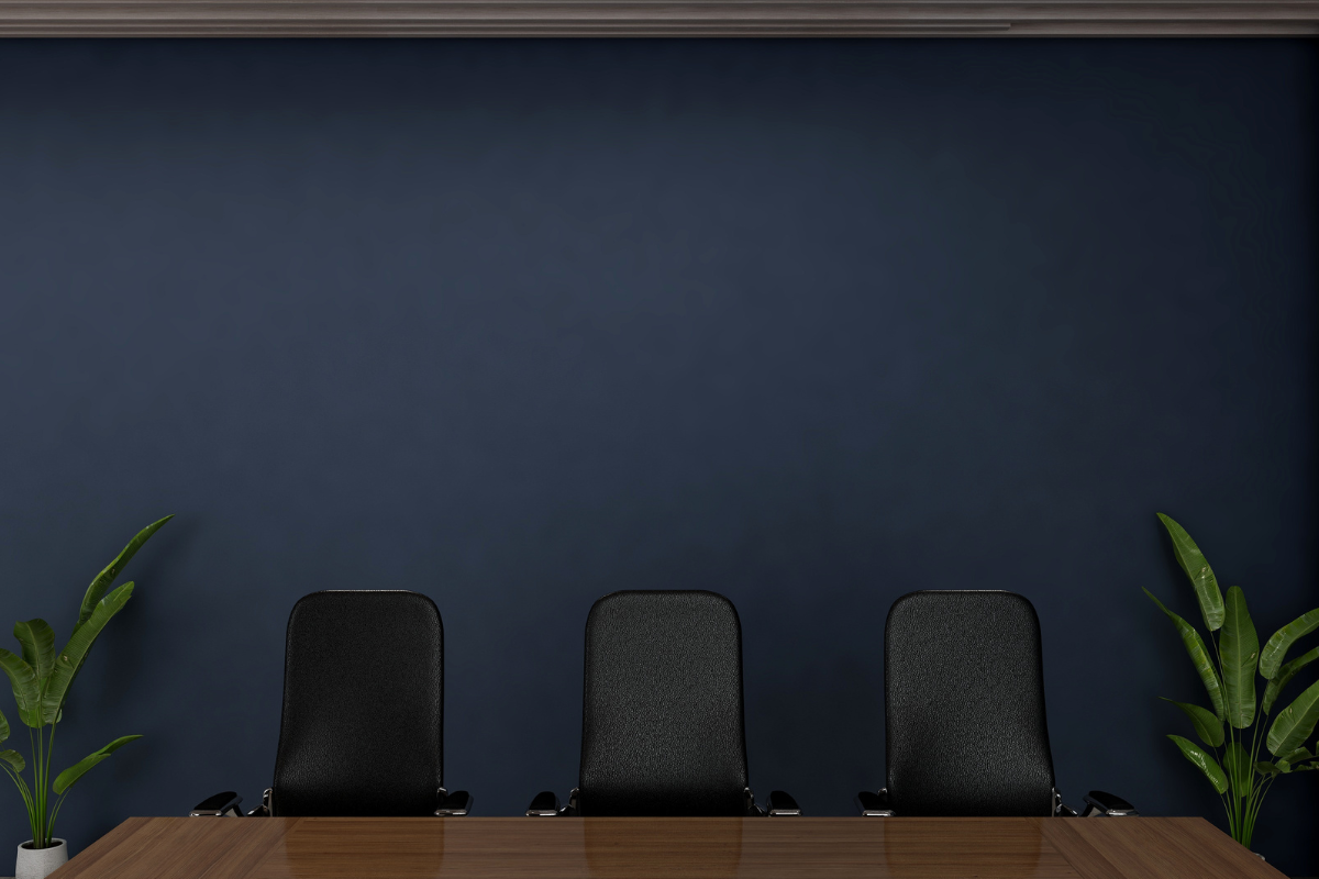 A modern office setup with a sleek dark blue painted wall, three black chairs, and wooden table, complemented by green plants on either side.
