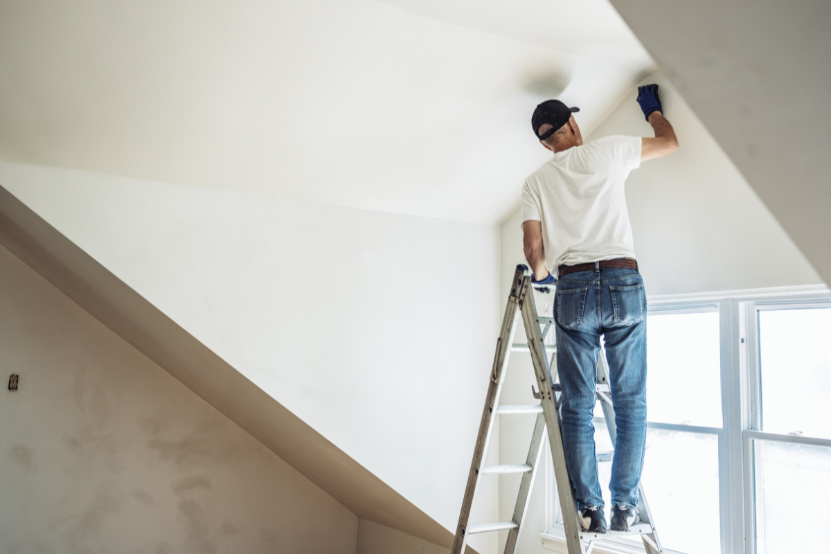 A painter on a ladder applies white paint to the ceiling of a bright, modern room.