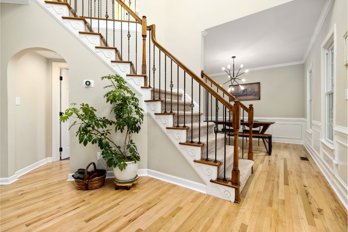 A beautifully designed staircase with white risers and beige carpeted treads, accented by dark wood handrails and black wrought iron balusters. The space includes hardwood flooring, a potted plant in a decorative pot, a wicker basket, and elegant lighting fixtures, creating a warm and inviting atmosphere.