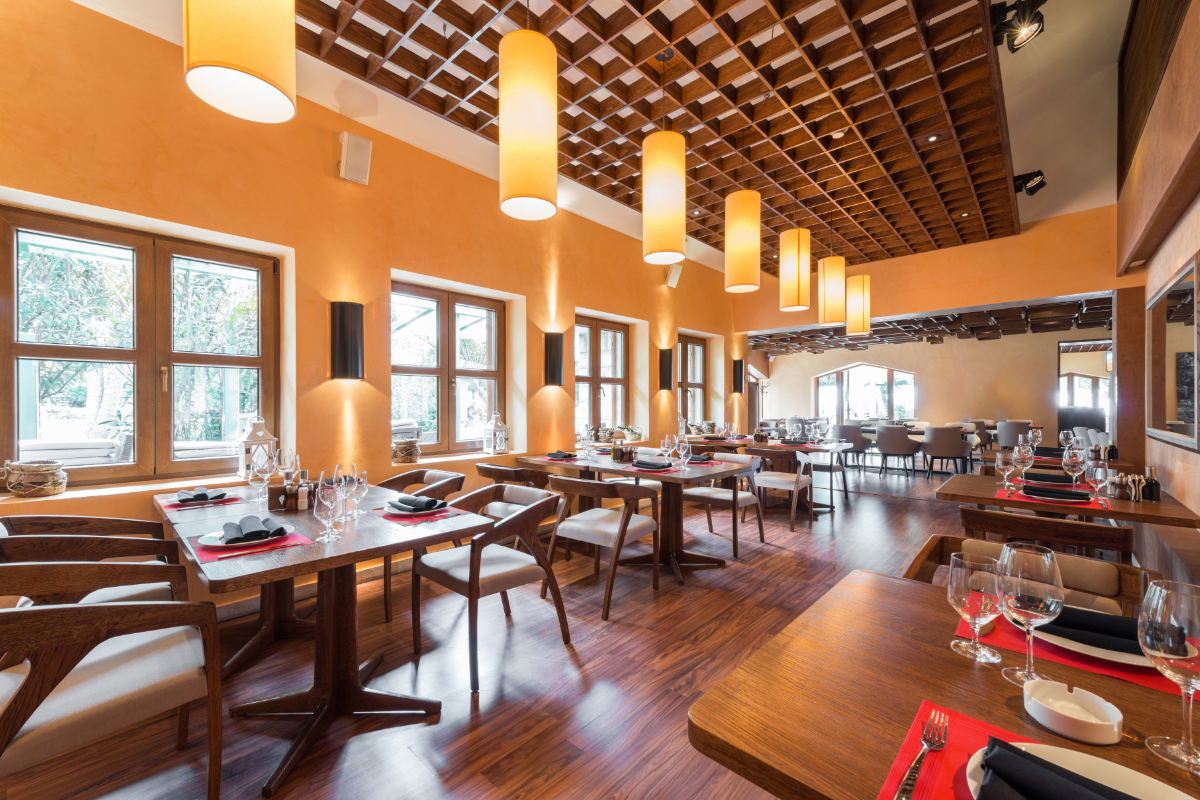 A warmly lit restaurant interior with orange-painted walls, wooden flooring, and hanging cylindrical lights, featuring neatly arranged tables and chairs.