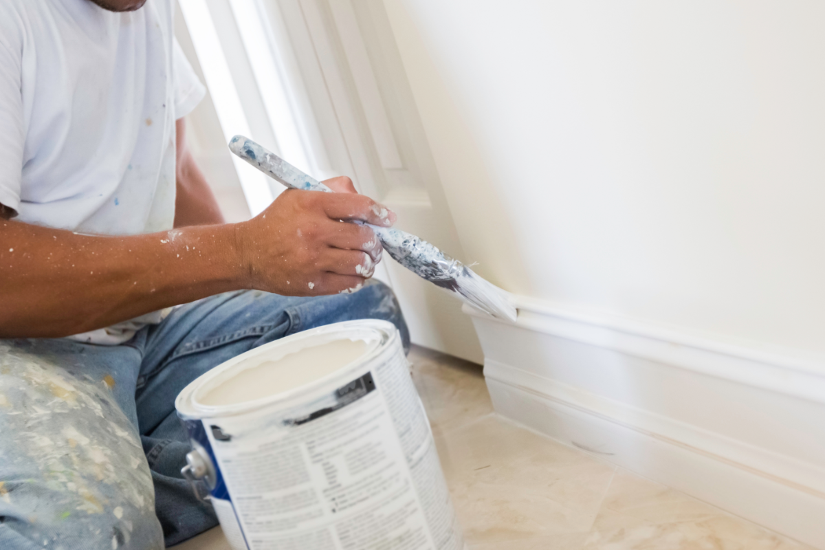 A painter is carefully applying white paint to the baseboard of a wall using a paintbrush. The individual, wearing a white shirt and blue jeans, is seated on the floor next to an open paint can, demonstrating precision in residential interior painting.