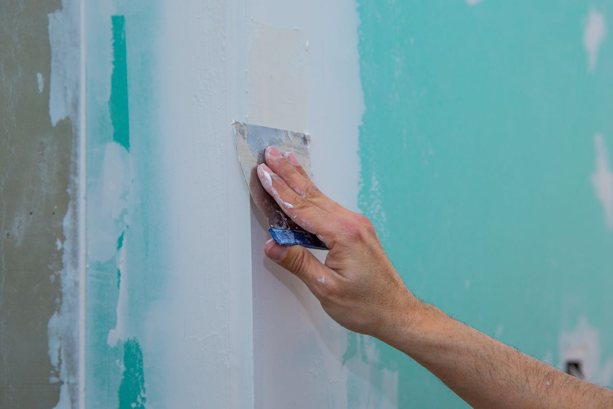 A person applying drywall compound to a wall using a putty knife, demonstrating the process of surface repair and preparation for painting.